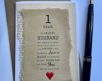 May include: A handmade anniversary card with the text "1 YEAR my AMAZING HUSBAND" on a textured paper background. A red heart is affixed to the card. A black fountain pen with a gold nib rests beside the card.