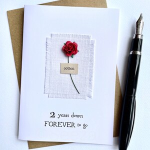 May include: A white greeting card with the text "2 years down FOREVER to go" and a red rose. The card features a small piece of white fabric with the word "cotton" on a label. A black fountain pen rests on the right side of the card.