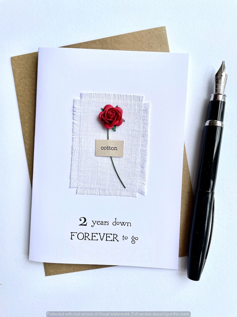 May include: A white greeting card with the text "2 years down FOREVER to go" and a red rose. The card features a small piece of white fabric with the word "cotton" on a label. A black fountain pen rests on the right side of the card.