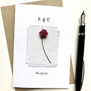 May include: A white greeting card with a burgundy rose and green stem, set against a textured white fabric. The card has the initials "R & E" and the date "26.09.22" printed on it. A black pen is placed to the right of the card.