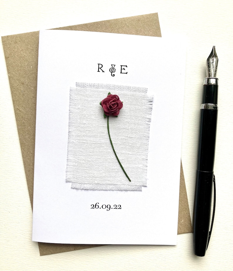 May include: A white greeting card with a burgundy rose and green stem, set against a textured white fabric. The card has the initials "R & E" and the date "26.09.22" printed on it. A black pen is placed to the right of the card.