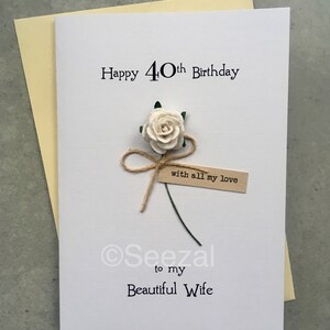 May include: A white birthday card with the text "Happy 40th Birthday" and "to my Beautiful Wife". It features a cream-colored paper rose, a twine bow, and a small tag that reads "with all my love". The card is placed on a light yellow envelope.