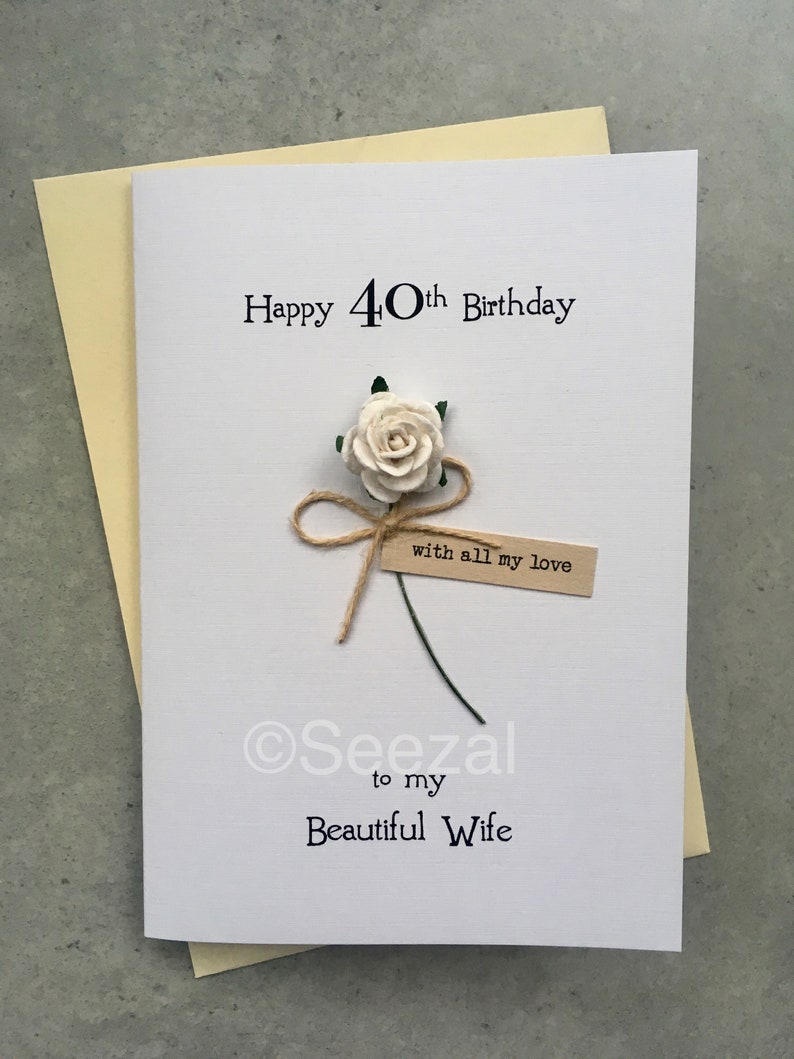 May include: A white birthday card with the text "Happy 40th Birthday" and "to my Beautiful Wife". It features a cream-colored paper rose, a twine bow, and a small tag that reads "with all my love". The card is placed on a light yellow envelope.