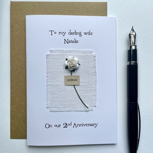 May include: A handmade anniversary card with the text "To my darling wife Natalie" and "On our 2nd Anniversary." The card features a white fabric square with a small white flower and the word "cotton." A black fountain pen rests beside the card.