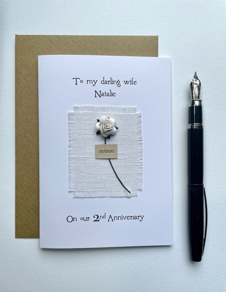 May include: A handmade anniversary card with the text "To my darling wife Natalie" and "On our 2nd Anniversary." The card features a white fabric square with a small white flower and the word "cotton." A black fountain pen rests beside the card.