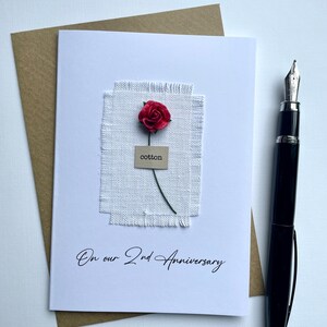 May include: A white card with a red paper rose and the word "cotton" on a small tag. The card reads "On our 2nd Anniversary" in script. A brown envelope and a black fountain pen are also present.