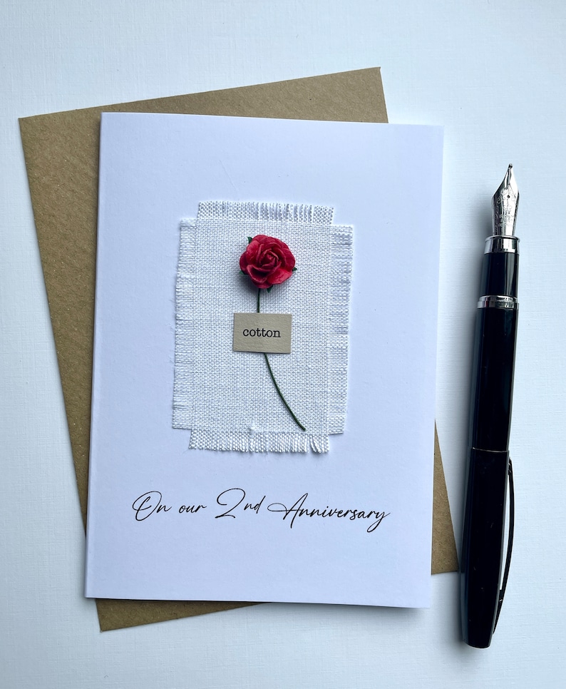 May include: A white card with a red paper rose and the word "cotton" on a small tag. The card reads "On our 2nd Anniversary" in script. A brown envelope and a black fountain pen are also present.