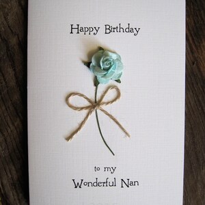 May include: A white birthday card with the text "Happy Birthday" and "to my Wonderful Nan". The card features a light blue paper flower with a twine bow and green stem.