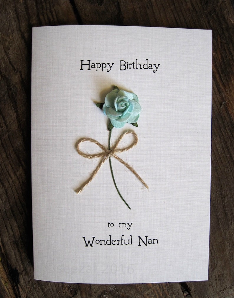May include: A white birthday card with the text "Happy Birthday" and "to my Wonderful Nan". The card features a light blue paper flower with a twine bow and green stem.