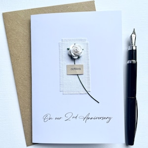 May include: A white card with the text "On our 2nd Anniversary" features a small white flower and the word "cotton." A brown envelope and a black pen are next to the card. The card has a rustic, handmade aesthetic.