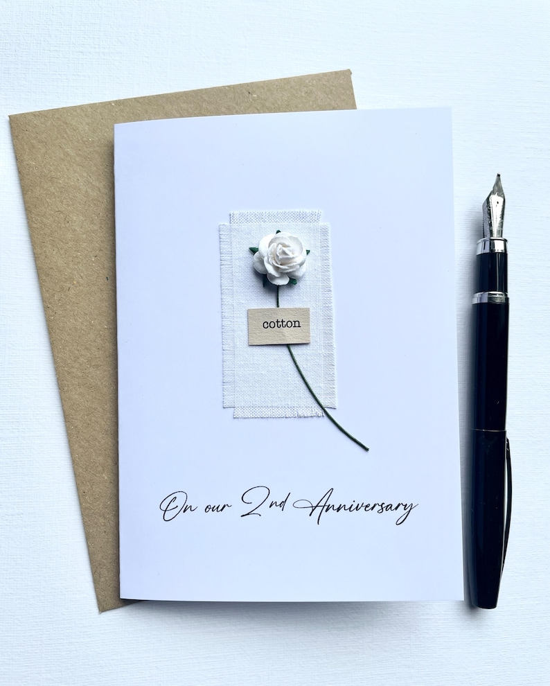 May include: A white card with the text "On our 2nd Anniversary" features a small white flower and the word "cotton." A brown envelope and a black pen are next to the card. The card has a rustic, handmade aesthetic.