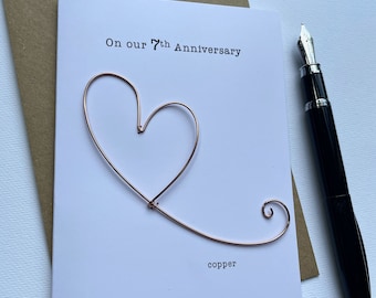 May include: A white anniversary card with the text "On our 7th Anniversary" and a copper wire heart design. The word "copper" is printed at the bottom. A black fountain pen rests on the side.