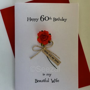 May include: A white greeting card with the text "Happy 60th Birthday" and "to my Beautiful Wife". It features a red rose with a small tag that reads "with all my love". The card is placed on a red envelope.