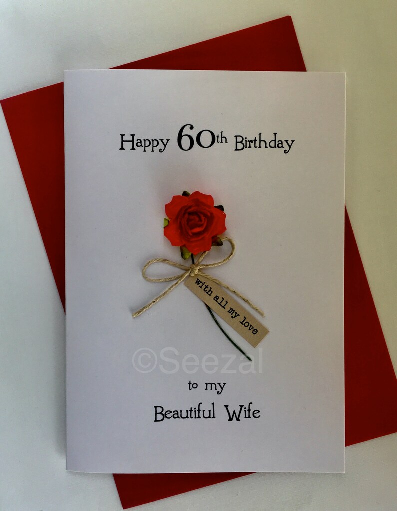 May include: A white greeting card with the text "Happy 60th Birthday" and "to my Beautiful Wife". It features a red rose with a small tag that reads "with all my love". The card is placed on a red envelope.