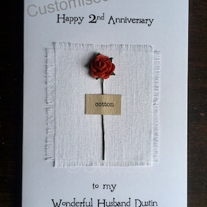 May include: A white greeting card with the text "Customised Happy 2nd Anniversary" and "to my Wonderful Husband Dustin". It features a small red rose, a square of white fabric, and a tag that says "cotton".