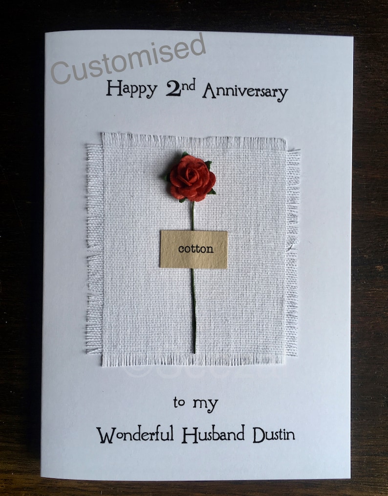 May include: A white greeting card with the text "Customised Happy 2nd Anniversary" and "to my Wonderful Husband Dustin". It features a small red rose, a square of white fabric, and a tag that says "cotton".