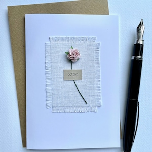 May include: A white greeting card with a light pink paper flower and the word "cotton" on a small tag. The card is on a white surface with a brown envelope and a black fountain pen.