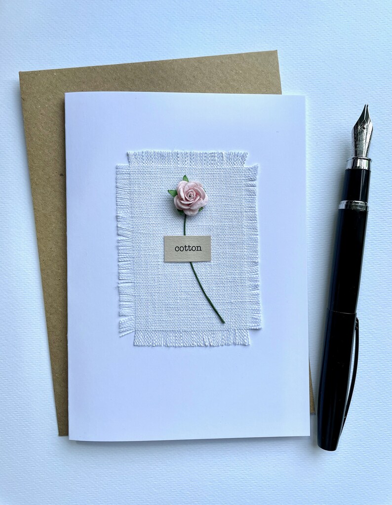 May include: A white greeting card with a light pink paper flower and the word "cotton" on a small tag. The card is on a white surface with a brown envelope and a black fountain pen.