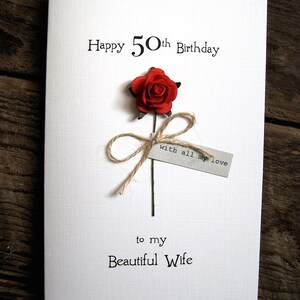 May include: A white greeting card with the text "Happy 50th Birthday" and "to my Beautiful Wife". It features a red paper rose with a twine bow and a small tag that reads "with all my love".