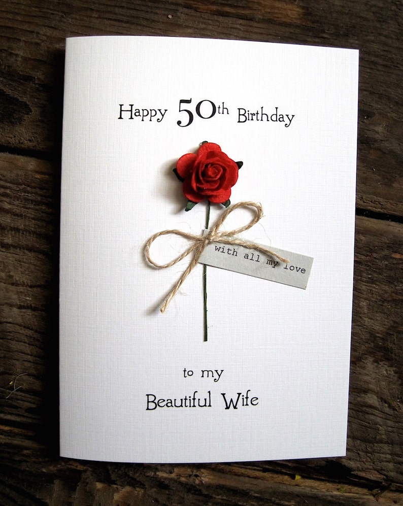 May include: A white greeting card with the text "Happy 50th Birthday" and "to my Beautiful Wife". It features a red paper rose with a twine bow and a small tag that reads "with all my love".