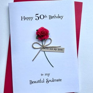May include: A white birthday card with the text "Happy 50th Birthday" and "to my Beautiful Soulmate". The card features a red paper rose with a twine bow and a small tag that reads "with all my love". The card is set against a red envelope.