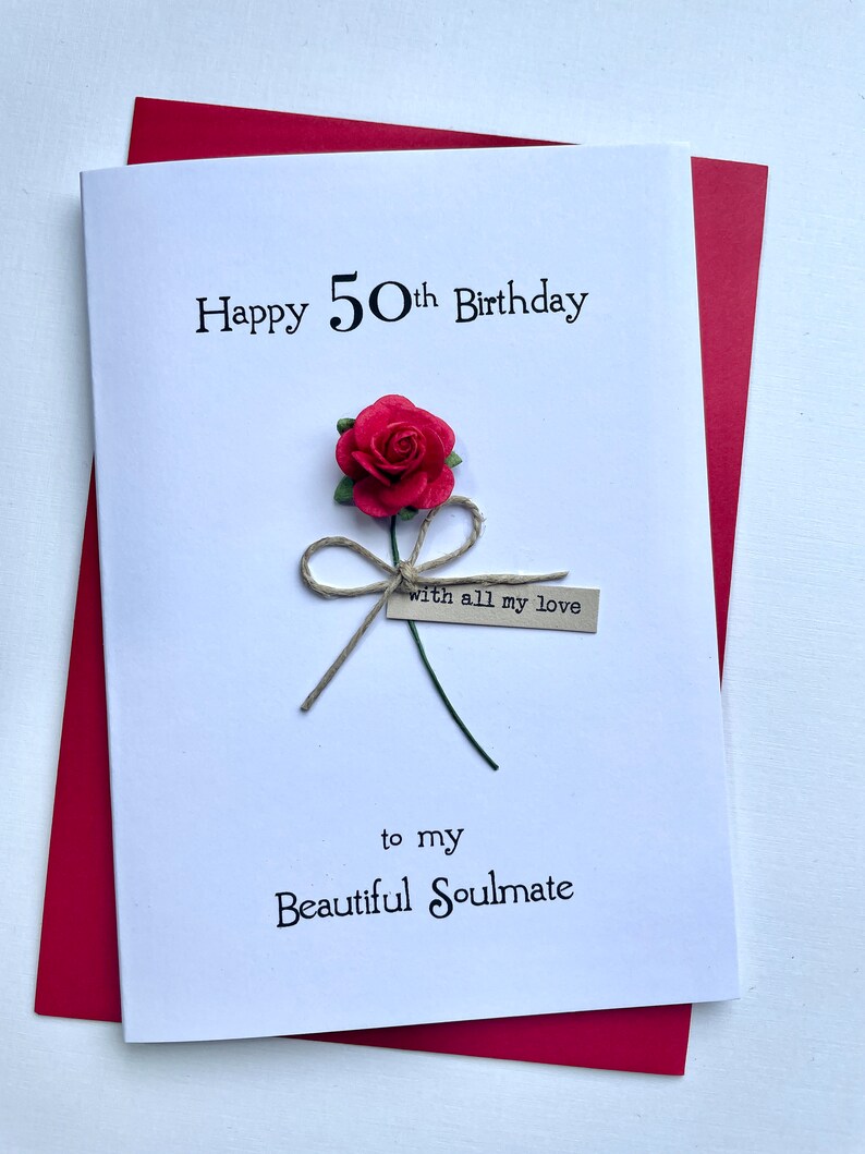 May include: A white birthday card with the text "Happy 50th Birthday" and "to my Beautiful Soulmate". The card features a red paper rose with a twine bow and a small tag that reads "with all my love". The card is set against a red envelope.