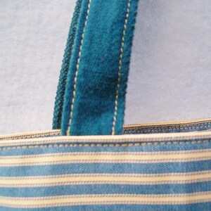 May include: Close-up of a blue and white striped fabric tote bag with a teal blue velvet strap. The strap has a gold thread running along the edge.