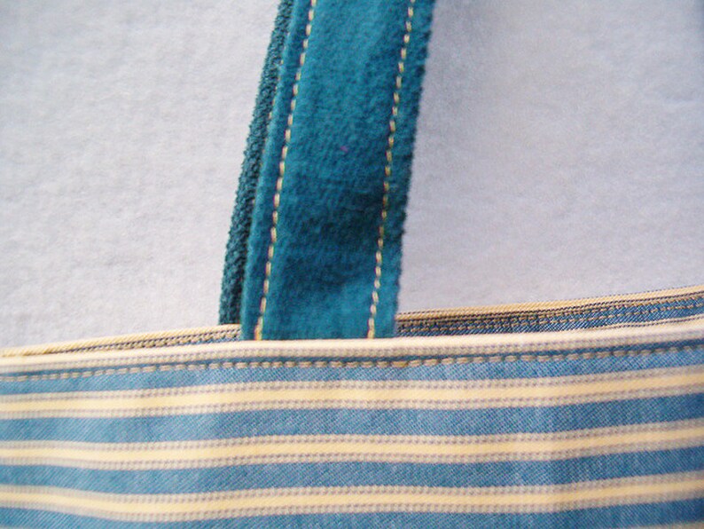 May include: Close-up of a blue and white striped fabric tote bag with a teal blue velvet strap. The strap has a gold thread running along the edge.