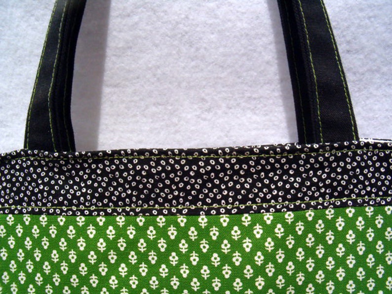 May include: A black and green tote bag with a white polka dot pattern on the top section and a smaller white floral pattern on the bottom section. The bag has black straps with green stitching.