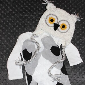 K&ouml;nnte beinhalten: Wei&szlig;er Baby-Strampler mit einem grauen und schwarzen Eulen-Design. Der Strampler hat eine passende geh&auml;kelte Eulenm&uuml;tze mit gelben Augen und einem schwarzen Schnabel.