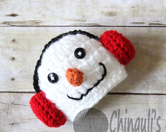 Puede incluir: Sombrero de ganchillo blanco en forma de muñeco de nieve con ojos negros, nariz naranja y una sonrisa cosida negra. El sombrero tiene orejeras de ganchillo rojas.