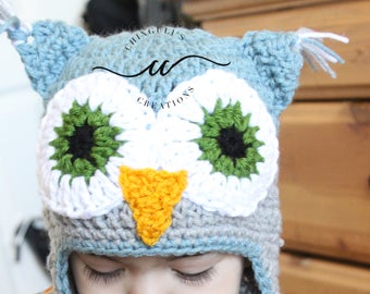 May include: A child wearing a gray and blue crocheted owl hat with white and blue braided ties. The hat has green and black eyes and a yellow beak.