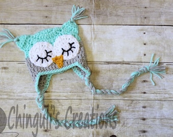 May include: A crocheted owl hat with grey and teal blue yarn. The hat has a long braided tail with tassels at the end. The owl's eyes are closed and it has a small orange beak.