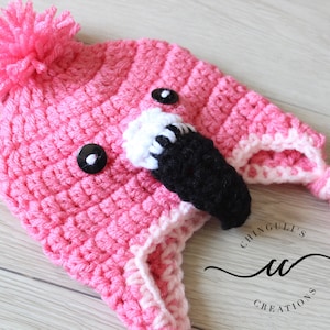 Könnte beinhalten: Rosa gestrickte Flamingo-Mütze mit schwarzen Augen, einem weiß-schwarzen Schnabel und einem rosa Bommel oben. Die Mütze hat einen langen rosa und weißen geflochtenen Schwanz.