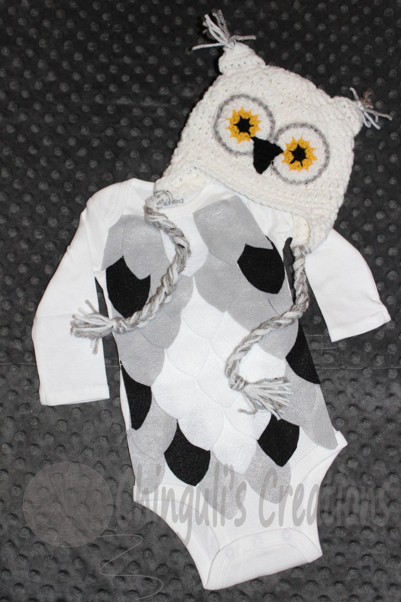 K&ouml;nnte beinhalten: Wei&szlig;er Baby-Strampler mit einem grauen und schwarzen Eulen-Design. Der Strampler hat eine passende geh&auml;kelte Eulenm&uuml;tze mit gelben Augen und einem schwarzen Schnabel.