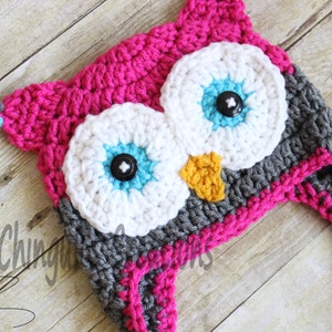 May include: A pink, gray, white, and blue crocheted owl hat with a braided tail. The hat has two large white eyes with blue and turquoise accents and black buttons for pupils.