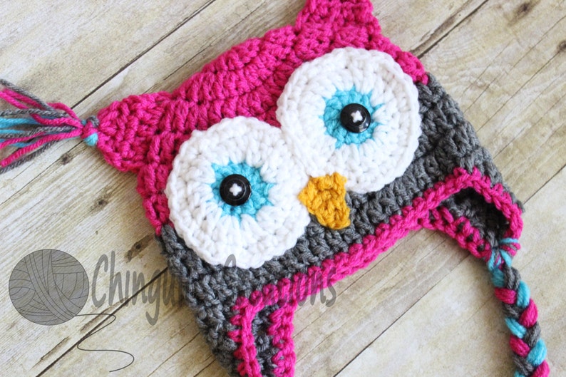 May include: A pink, gray, white, and blue crocheted owl hat with a braided tail. The hat has two large white eyes with blue and turquoise accents and black buttons for pupils.
