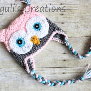 May include: A pink, gray, and blue crocheted owl hat with a long braided tail. The hat has large white eyes with blue and black accents and a yellow beak.