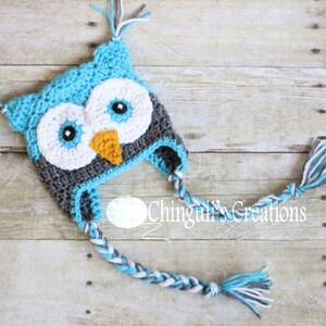 May include: A blue, gray, and white crocheted owl hat with long, braided ties. The hat has a large, round face with black eyes and an orange beak.