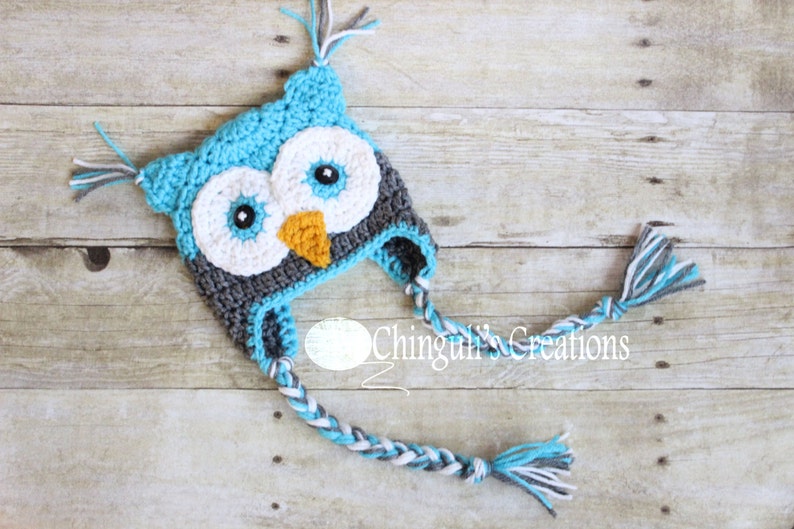 May include: A blue, gray, and white crocheted owl hat with long, braided ties. The hat has a large, round face with black eyes and an orange beak.