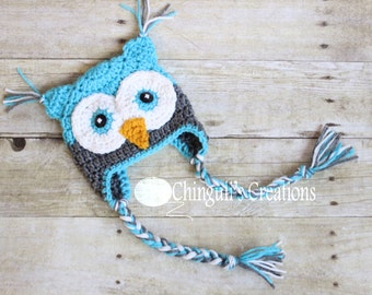May include: A blue, gray, and white crocheted owl hat with long, braided ties. The hat has a large, round face with black eyes and an orange beak.