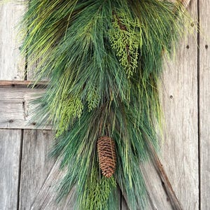 May include: A teardrop-shaped evergreen swag featuring long, slender green pine needles mixed with flat, textured cedar foliage. The arrangement is accented with several brown pinecones distributed throughout the greenery, creating a natural look.