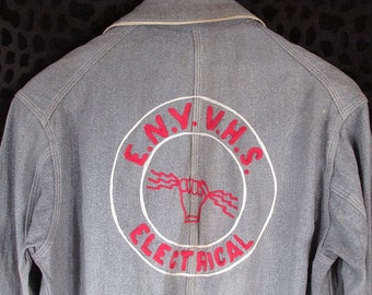 Può includere: Una giacca da lavoro grigia con una toppa ricamata in rosso e bianco sul retro. La toppa è un cerchio con il testo "E.N.V. V.H.S. ELECTRICAL" e un'immagine stilizzata di una mano che tiene un filo.