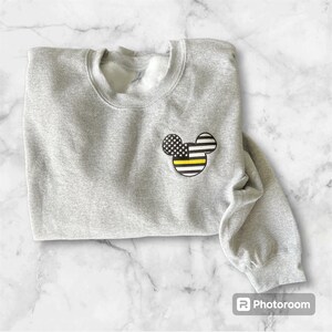 May include: A gray sweatshirt with a black and white American flag Mickey Mouse design with a yellow stripe across the middle. The flag is a thin yellow line flag.