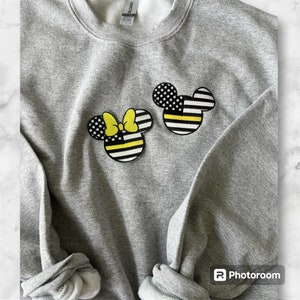 May include: Gray sweatshirt with two embroidered patches of Mickey Mouse and Minnie Mouse. The patches are in the style of the American flag with a yellow and black thin line representing the "Thin Yellow Line" for law enforcement.