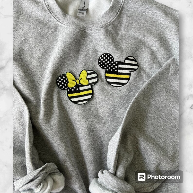 May include: Gray sweatshirt with two embroidered patches of Mickey Mouse and Minnie Mouse. The patches are in the style of the American flag with a yellow and black thin line representing the "Thin Yellow Line" for law enforcement.