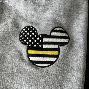 May include: A gray sweatshirt with a black and white embroidered Mickey Mouse design. The Mickey Mouse ears are shaped like the American flag with a yellow thin line across the middle.