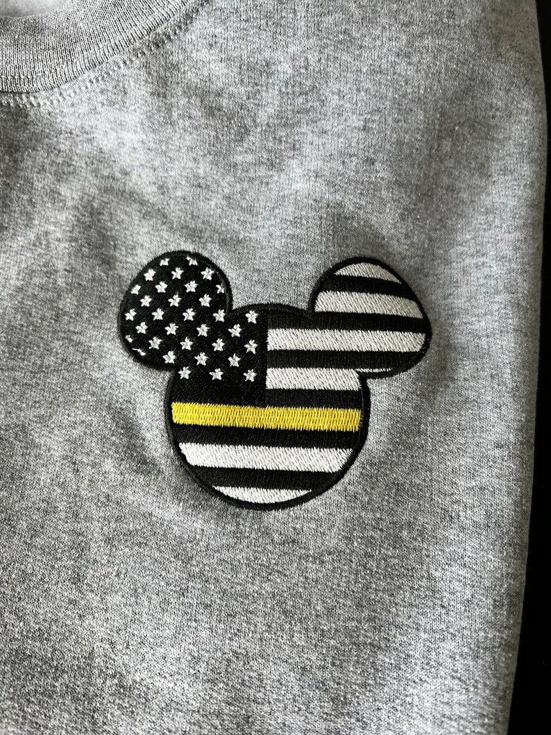 May include: A gray sweatshirt with a black and white embroidered Mickey Mouse design. The Mickey Mouse ears are shaped like the American flag with a yellow thin line across the middle.