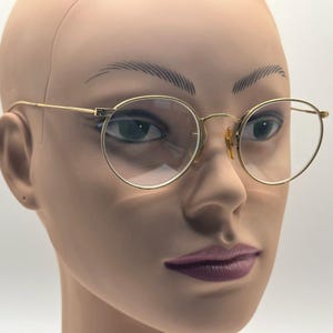 May include: Gold-rimmed eyeglasses with a classic round frame design. The glasses have a thin, delicate frame and clear lenses. The image shows the glasses on a mannequin head, highlighting the shape and style of the eyewear.