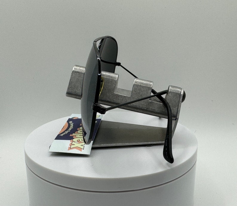 May include: Black sunglasses with a metal frame and black arms. The sunglasses are displayed on a silver stand with a white base.